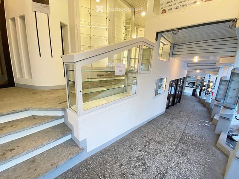 A wide, open corridor with white walls, glass display cases, and a polished stone floor. The corridor features a staircase leading upwards and several glass display units lining the walls.