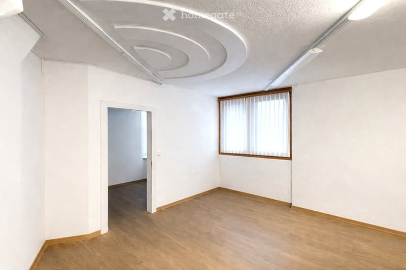 Empty room with white walls, wooden flooring, and a large window with white blinds. The ceiling features modern lighting fixtures.