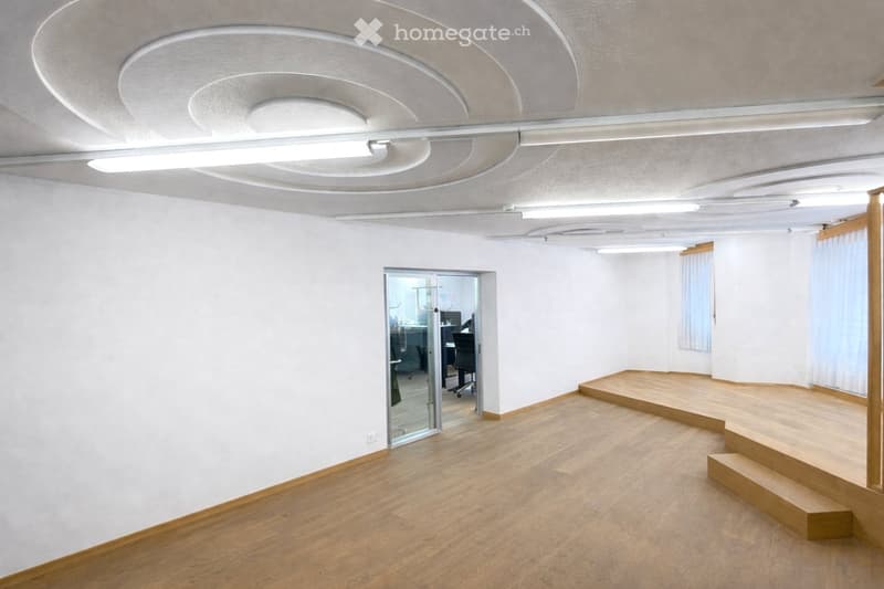 Empty room with light wooden flooring, white walls, and a raised platform area. A glass door leads to an adjacent room with office furniture visible.