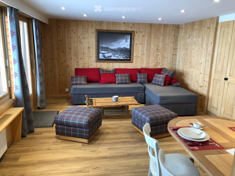 Cozy living room with wooden walls and floor, red sofa with plaid cushions, coffee table, ottomans, dining table with chairs, and a scenic mountain view picture on the wall.