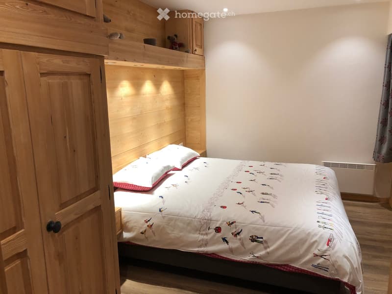 Cozy wooden bedroom with a double bed, pillows, a decorative quilt, a small lofted area with a teddy bear, and a wooden wardrobe.