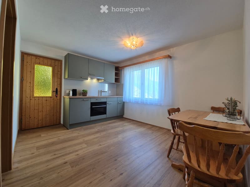 A modern kitchen and dining area with grey cabinets, a wooden dining table, and wooden chairs. The space features a wooden floor, a window with white curtains, and a small plant on the counter.