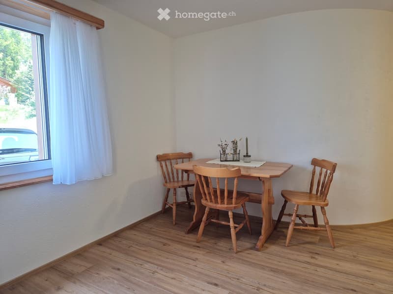 A small wooden dining area with a table, four chairs, a vase with flowers, and a candle on the table. The room has wooden flooring and white walls with a window on one side.