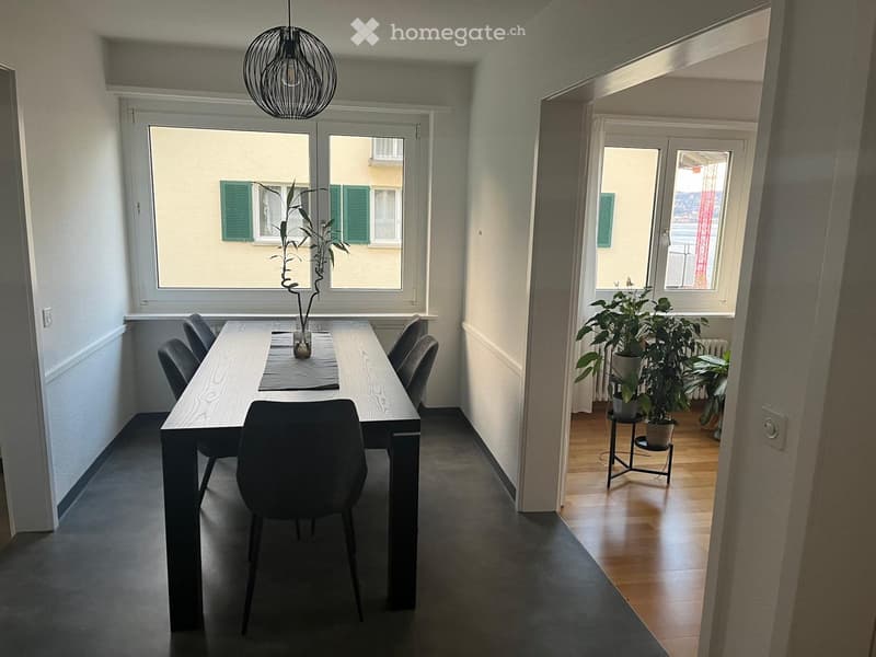 Dining area with a long wooden table, four chairs, and a potted plant on the table. The adjacent room has wooden flooring and additional potted plants.