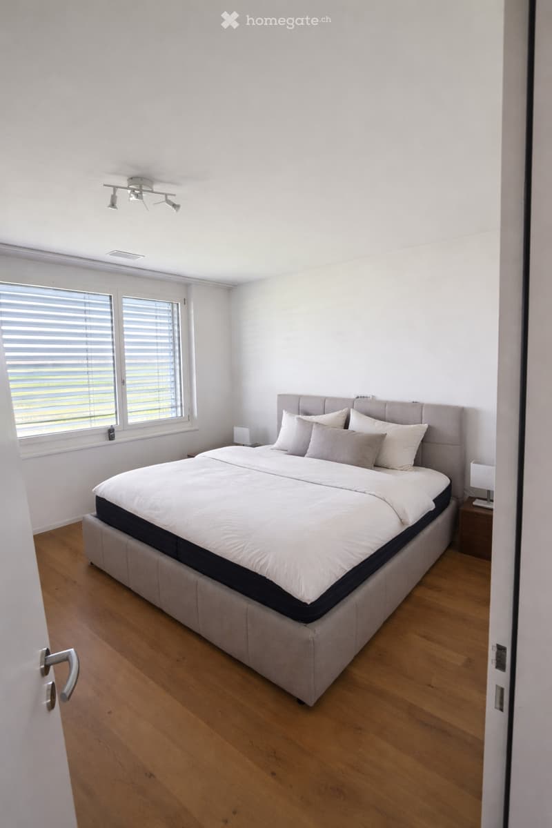 Modern bedroom with a large bed, white bedding, wooden floor, and a nightstand with a lamp. The room features large windows with blinds and recessed ceiling lighting.