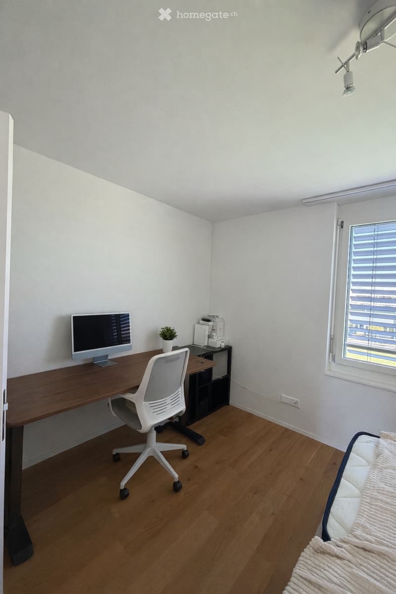 A modern, well-lit room with a wooden floor, a large desk with a computer monitor, a white office chair, a small plant, a printer, and a bed with a white blanket.