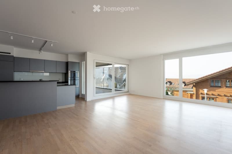 Bright, empty room with wooden flooring, large windows, and a modern kitchen area with grey cabinets and a countertop.