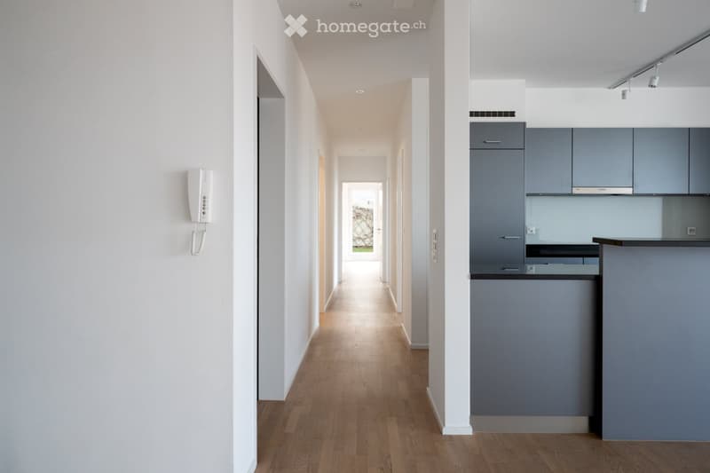 Modern hallway with light wooden flooring, white walls, and recessed ceiling lighting. A small kitchen area with grey cabinets and a countertop is visible on the right side.
