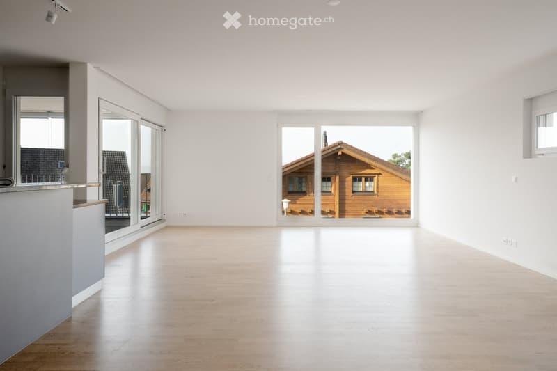 Bright, empty room with light wooden flooring and white walls. Large windows and sliding glass doors offer views of a wooden house outside. Minimalist design with no furniture.