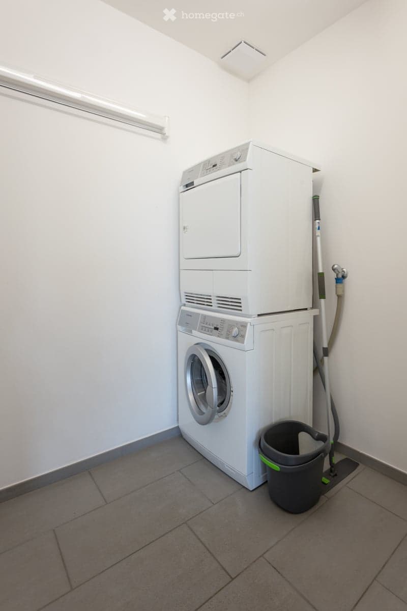 A small, clean laundry room with a stacked washing machine and dryer, a broom, and a bucket on the floor.