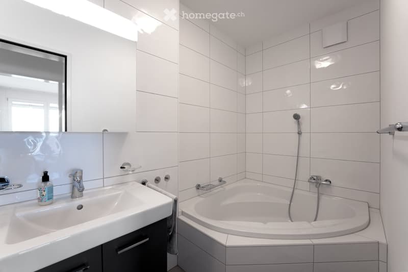 Modern bathroom with white tiled walls and floor, featuring a corner bathtub, a sink with a modern faucet, and a large mirror above it. A soap dispenser is placed on the sink counter.