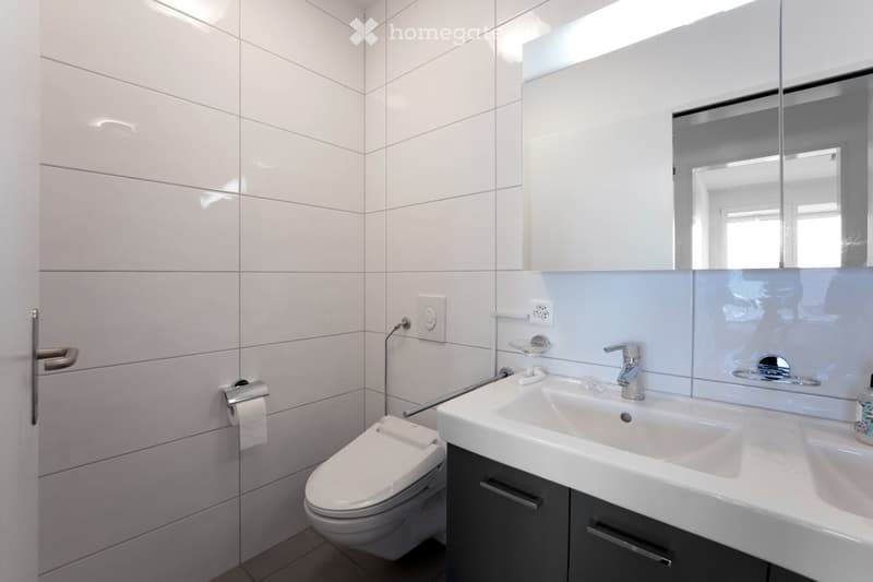 Modern bathroom with white tiles, a toilet, a double sink with a chrome faucet, a large mirror, and a dark grey cabinet.