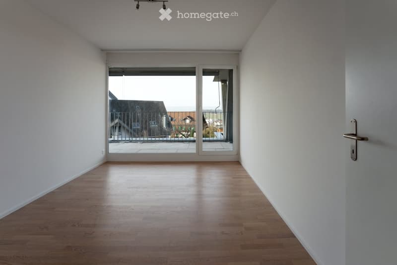 Empty room with wooden flooring, white walls, and large sliding glass doors leading to a balcony with a view of neighboring houses.