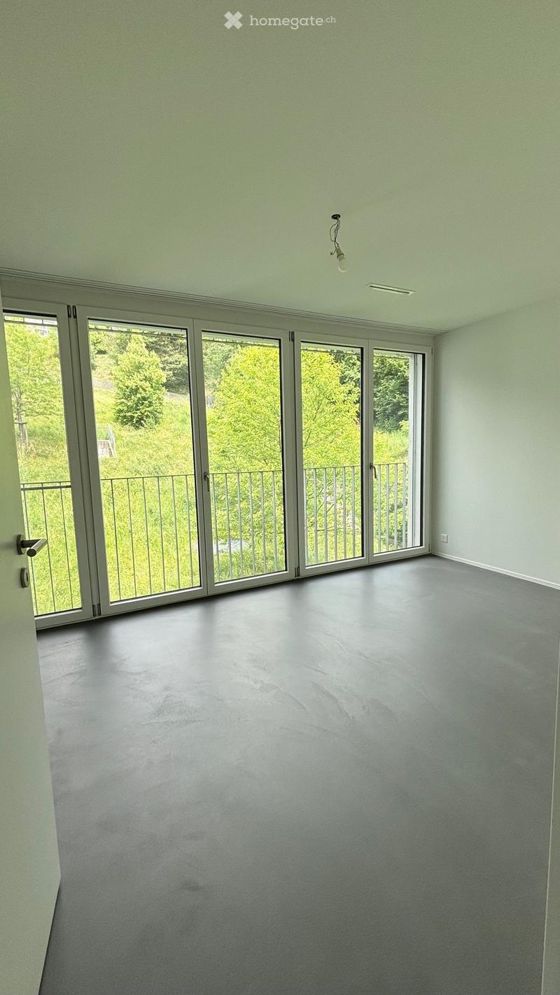 Empty room with light green walls, grey floor, large sliding glass doors leading to a balcony with a metal railing, and a single light fixture on the ceiling.