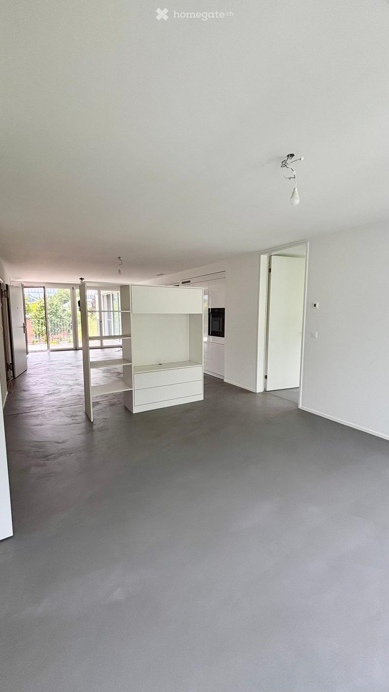 Empty room with grey carpeted floor, white walls, and a single white shelving unit. The room has large windows and doors leading to an outdoor area.