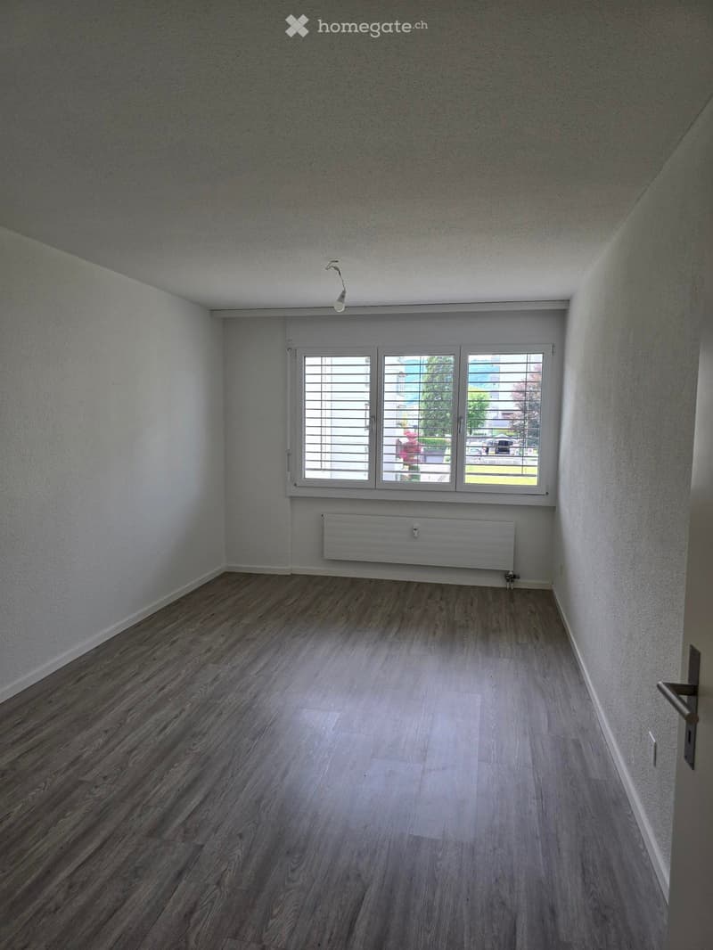 Empty room with wooden flooring, white walls, and a large window with blinds. A radiator is mounted on the wall below the window.