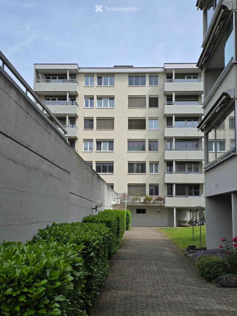 Modern multi-story residential building with balconies and large windows, surrounded by trimmed hedges and a paved walkway.