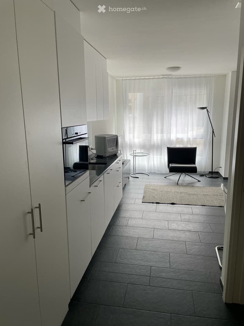 Furnished Room. A modern, minimalist room with a dark tiled floor, white cabinets, a television on a counter, a small round table, a single chair, and a floor lamp near a large window with sheer curtains.