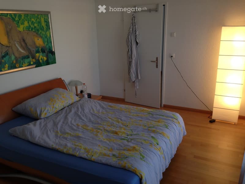Bedroom with a single bed, nightstand, lamp, and a picture frame on the wall. Wooden flooring and a door with a striped towel hanging on it.