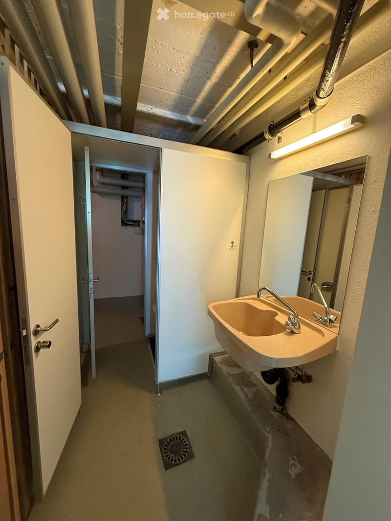A narrow hallway with exposed pipes and a double sink in the corner, leading to another room.