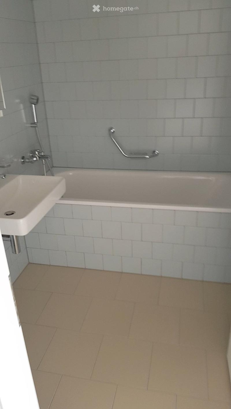 Bathroom with a bathtub, sink, and handrail. Light blue tiles on walls and a beige tiled floor.