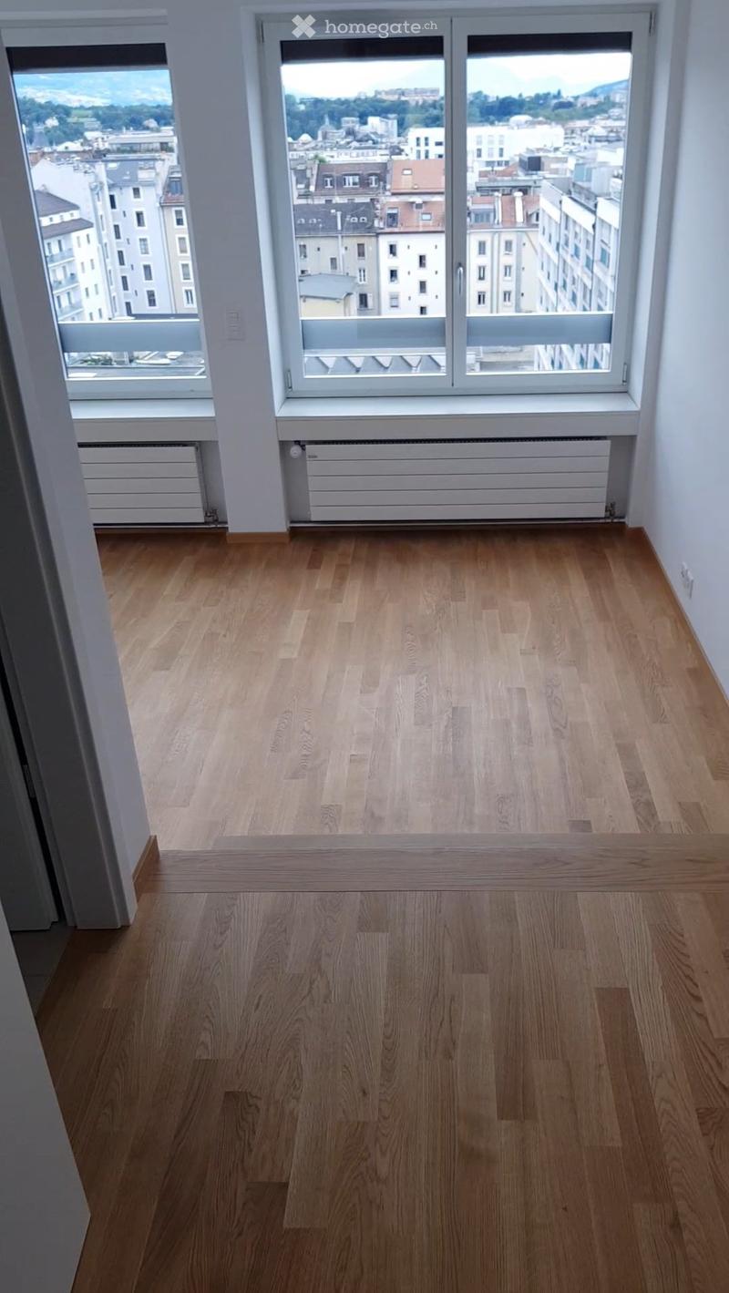Empty room with light wooden flooring, large windows offering a cityscape view, and white walls.