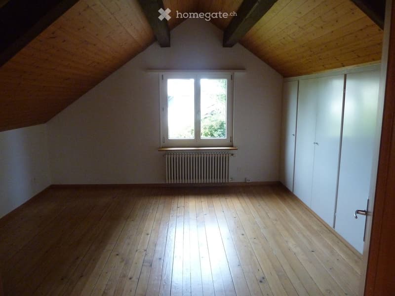 Empty attic room with wooden floors, white walls, and a slanted wooden ceiling. Features a window with white frames and a radiator below it.