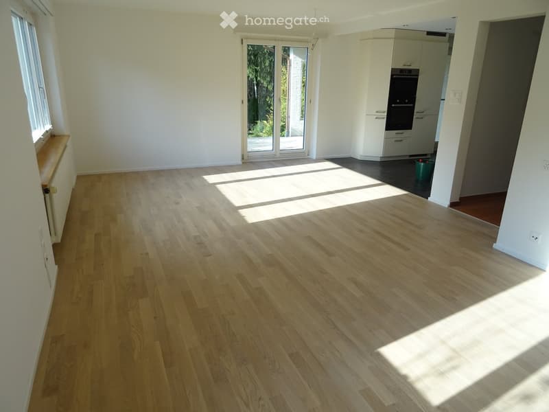 Empty room with light wooden flooring, white walls, large windows, and a sliding glass door leading to an outdoor area. The adjacent kitchen area is visible with cabinets and an oven.