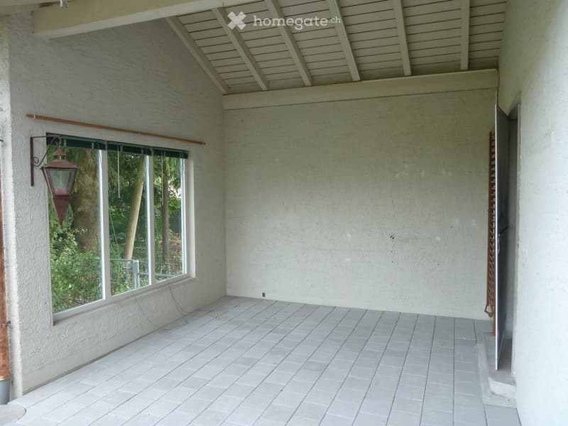 Empty balcony with white tiled floor, white walls, large window with view of greenery, and a decorative lantern-style light fixture on the left.