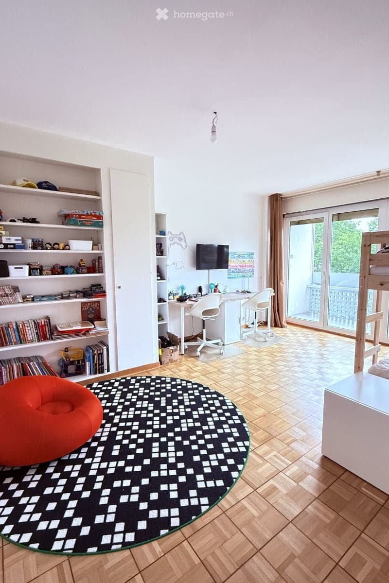 A modern, well-lit room with a wooden floor, large window, and a mix of furniture including a sofa, table, chair, shelves, and a screen.