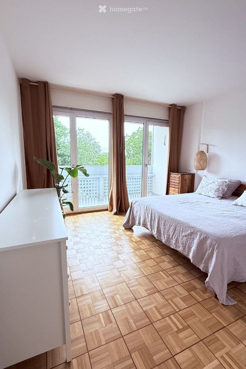 Bright bedroom with wooden herringbone flooring, large sliding glass doors leading to a balcony, a neatly made bed with pillows and a duvet, a nightstand, and a potted plant near the window.