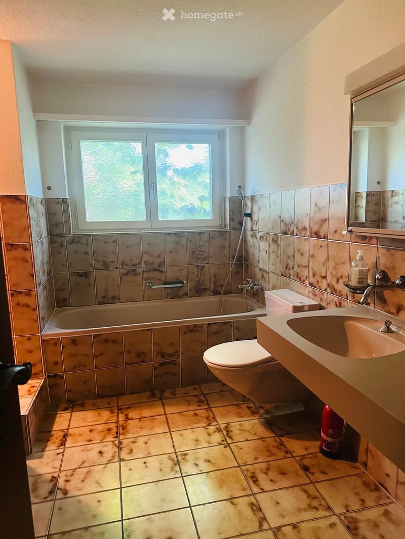 Bathroom with a bathtub, toilet, sink, mirror, and a window with frosted glass. The walls are tiled with a mix of brown and beige tiles, and the floor is tiled with a light brown tile pattern.