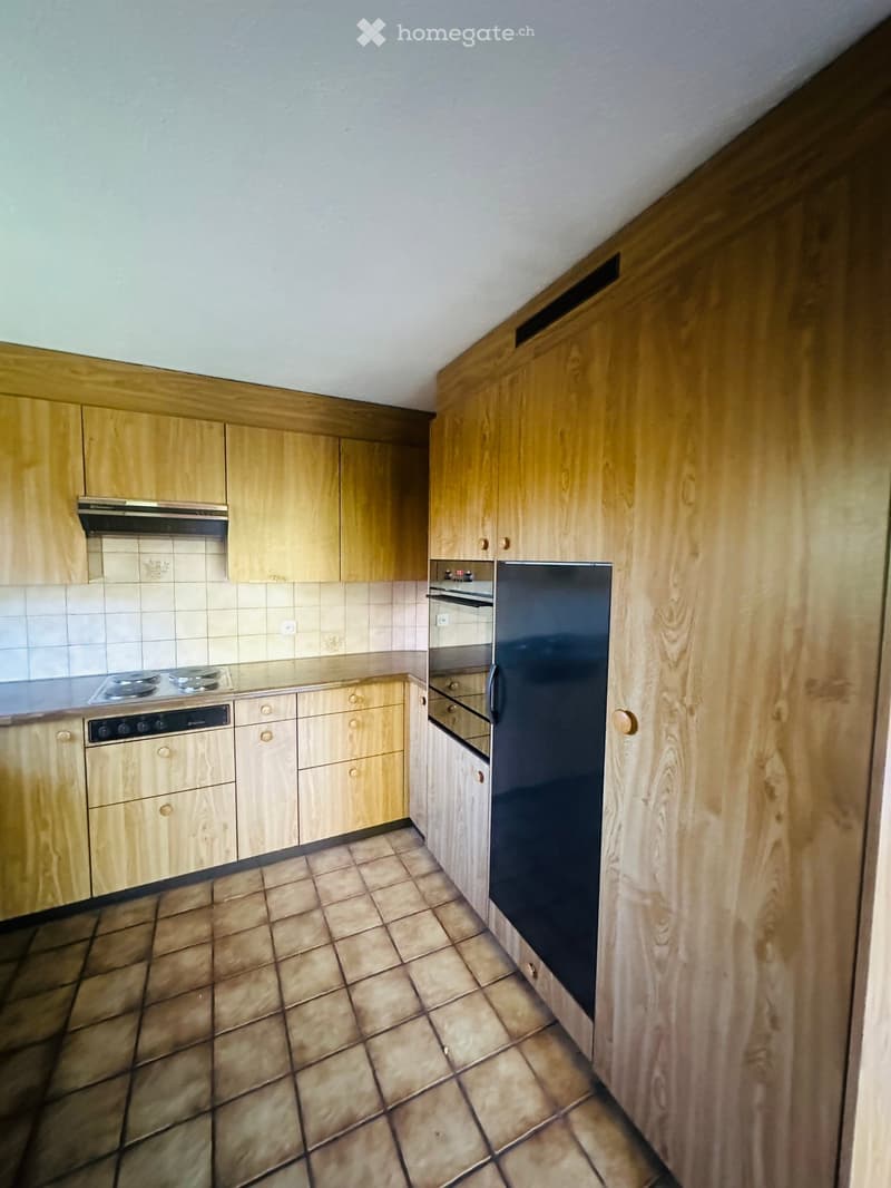 A small kitchen with light wooden cabinets, a black refrigerator, a stove, and tiled flooring.