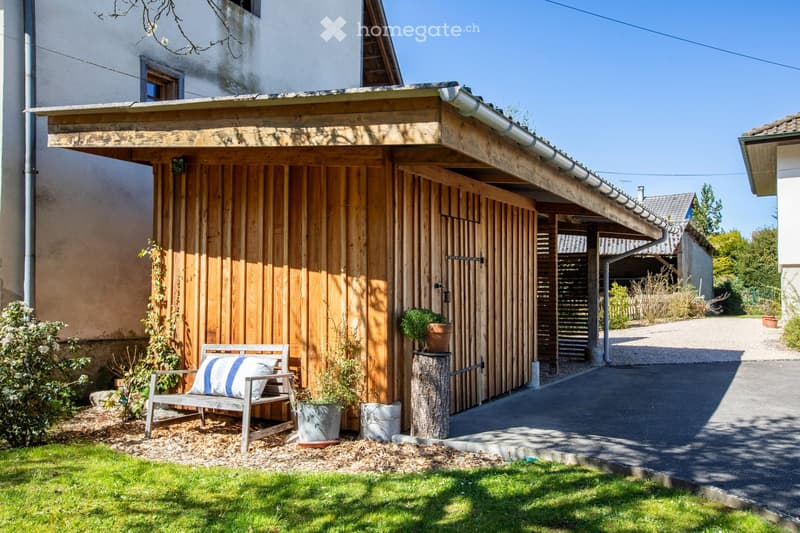 Wooden garage with a bench and potted plants, driveway, and a house in the background