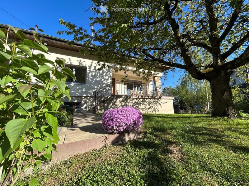 Two-story house with a balcony, surrounded by a well-maintained garden with a large tree and flowering bush.