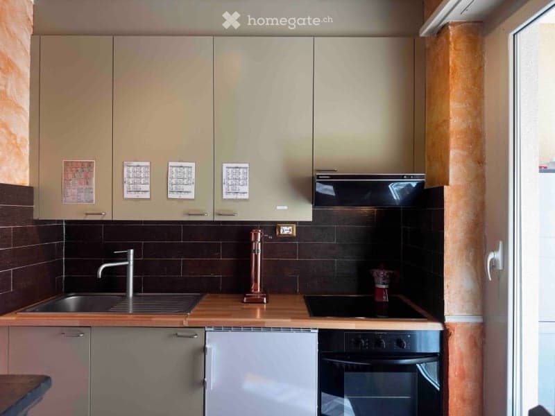 Modern kitchen with light-colored cabinets, dark tiled backsplash, and stainless steel appliances including a sink, stove, and oven. A decorative copper faucet is visible on the countertop.