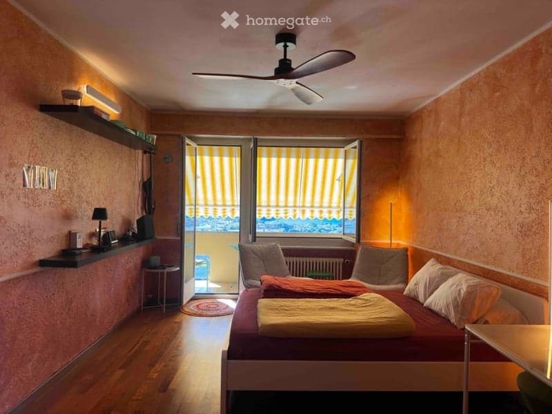 Cozy bedroom with wooden flooring, a double bed with pillows and blankets, a nightstand with a lamp, a shelf with various items, and an open door leading to a balcony with yellow blinds.