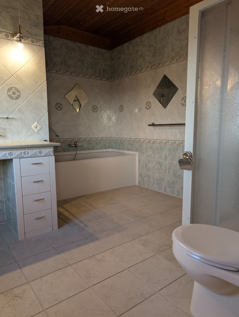 Bathroom with a bathtub, toilet, vanity, and mirror. The walls are tiled with a marble pattern, and the floor is tiled with large beige tiles.