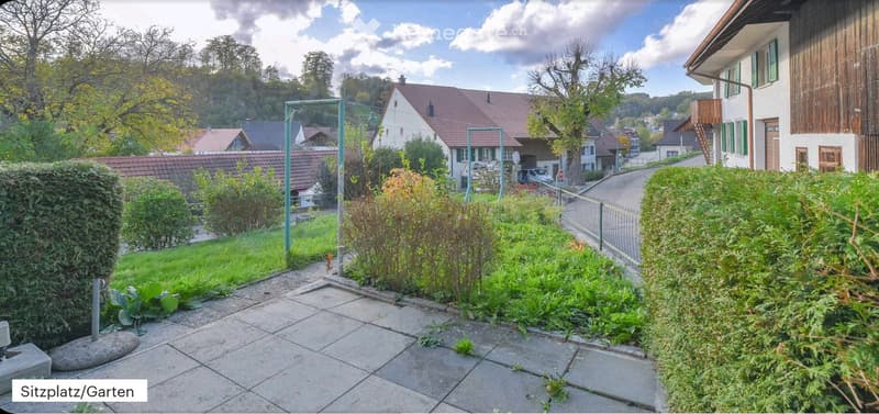 Garden area with paved patio, greenery, and surrounding houses visible in the background.