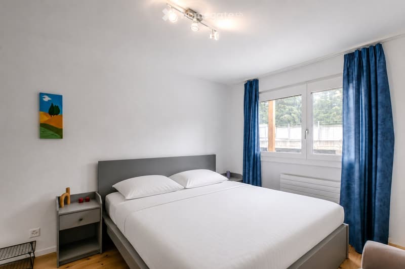 Modern bedroom with a double bed, nightstand, and large window with blue curtains. Features a painting on the wall and a radiator below the window.