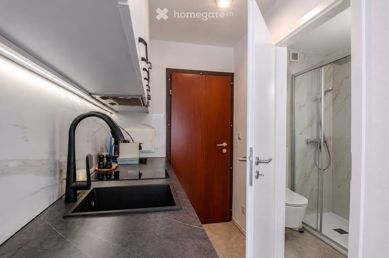 Modern bathroom with a sleek black faucet, a large sink, a glass-enclosed shower, and a toilet. The space features marble-like tiles and a wooden door leading to another room.