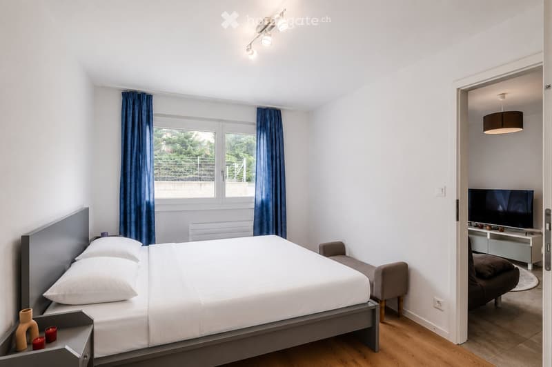Modern bedroom with a double bed, nightstand, chair, and a small living area with a TV and sofa.