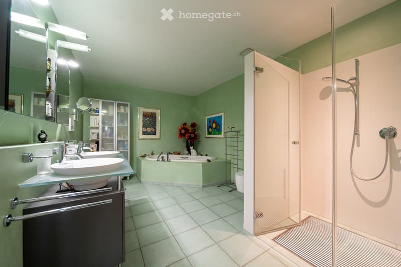 Modern bathroom with green walls, white tiled floor, double sink vanity, bathtub, separate shower, and decorative elements including framed artwork and a flower arrangement.