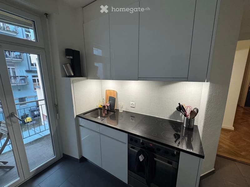 Modern kitchen with grey cabinets, stainless steel appliances, and a balcony view. Includes a sink, oven, cutting board, and various kitchen utensils.
