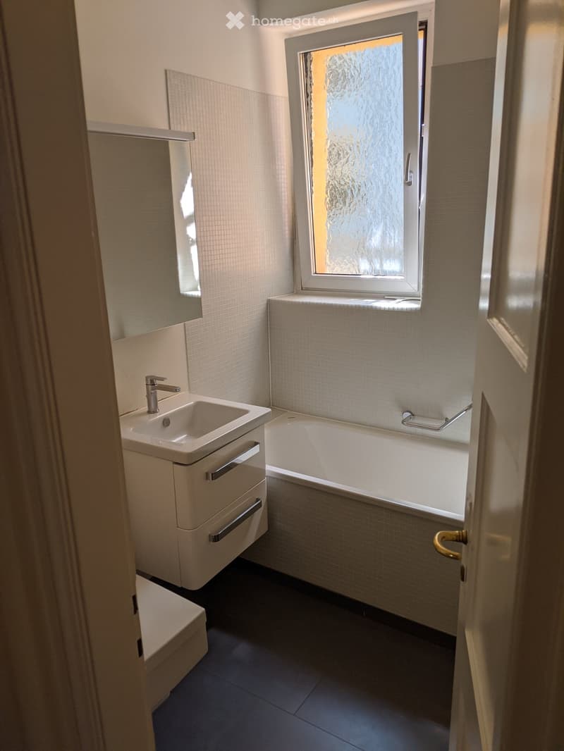 Modern bathroom with a white sink, bathtub, and frosted glass window. The walls are tiled, and the floor is dark.