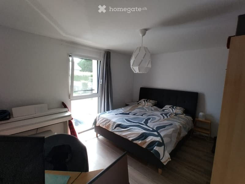 Bright bedroom with a large bed, nightstand, chair, and a window with curtains. The room features a modern lamp and a small table with a chair.