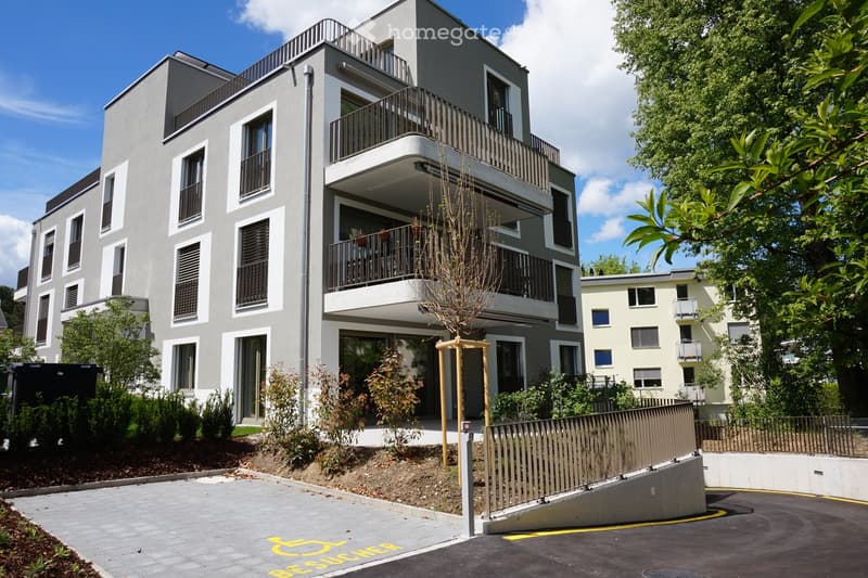 Modern multi-story residential building with balconies, large windows, and contemporary design. The building features a raised ground floor and is surrounded by greenery and a paved parking area.
