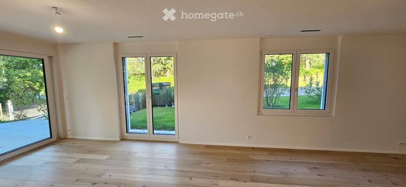 Empty room with light wooden flooring, white walls, large windows, and sliding glass doors leading to a green outdoor area.