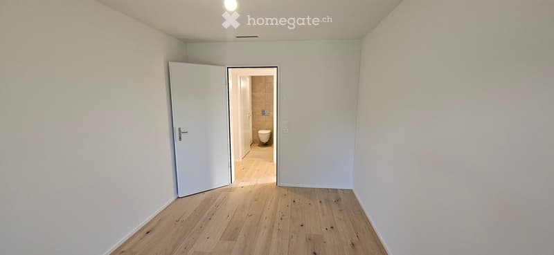 Empty hallway with light wooden flooring and white walls, leading to a small bathroom with a toilet and tiled walls.