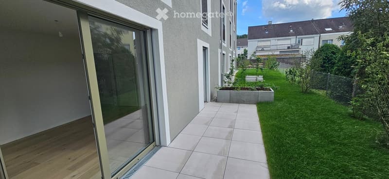 Modern outdoor patio area with tiled flooring, glass sliding door leading to interior, small garden with grass, plants, and a raised planter box.